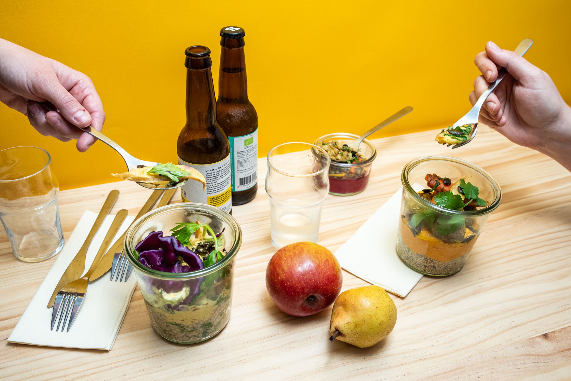 Deux mains tiennent chacune une fourchette prête à déguster des bocaux de nourriture entouré de bouteille en verre et de fruits sur une table en bois et avec un fond jaune.